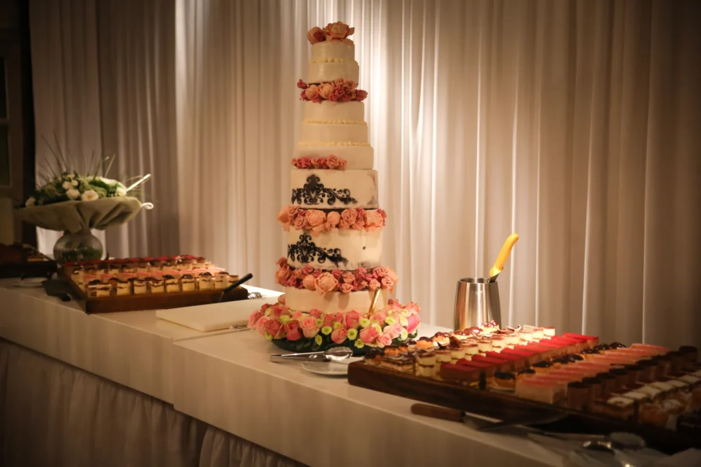 Hoge witte bruidstaart met lagen roze rozen en zwarte versieringen op een buffet met gebakjes