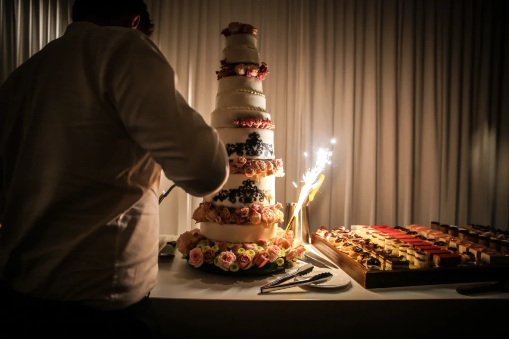 Man snijdt een hoge witte bruidstaart met roze rozen aan, met sterretjesvuurwerk ernaast en een plank met gebakjes